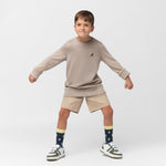 A young boy with short brown hair strikes a playful pose, standing with his legs apart and arms bent outward. He wears a beige oversized long-sleeve sweatshirt with a small embroidered brown dog logo on the left chest, beige shorts, navy blue socks with a yellow pineapple pattern, and white sneakers with green and brown accents. The background is plain white.