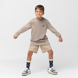 A young boy with short brown hair strikes a playful pose, standing with his legs apart and arms bent outward. He wears a beige oversized long-sleeve sweatshirt with a small embroidered brown dog logo on the left chest, beige shorts, navy blue socks with a yellow pineapple pattern, and white sneakers with green and brown accents. The background is plain white.