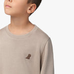 A close-up of a young boy wearing a beige long-sleeve sweatshirt with a ribbed crew neckline. The focus is on the small embroidered brown dog logo on the left chest area. The fabric has a subtle texture, and the stitching details are visible. The boy's face is partially visible, showing his chin and lips as he looks slightly upward. The background is plain white