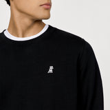 A close-up of a black crewneck sweatshirt featuring a small embroidered logo on the left chest. The logo depicts a playful dog with a unique design. The model is wearing a white shirt underneath, visible at the neckline, adding a layered look to the outfit.