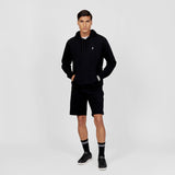 Full-body view of a man wearing a black hoodie paired with black shorts. The hoodie features a small embroidered white dog logo on the chest and includes drawstrings. The outfit is styled with black crew socks with white stripes and black sneakers, layered over a white t-shirt