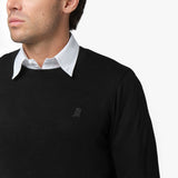  A close-up of a man wearing a black crewneck sweatshirt over a white collared shirt. The image focuses on the upper chest area, highlighting a small embroidered logo on the left side of the sweatshirt, which features a stylized dog. The black-on-black embroidery blends subtly with the fabric. The man’s face is partially visible, showing his jawline and lips, while he maintains a neutral expression against a plain white background