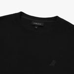 A close-up of a black crewneck sweatshirt, showing the ribbed collar and finely knit fabric texture. The black neck label displays the brand name "JAMES BARK" with "Est. 2015" beneath it, along with the size "M." A subtly embroidered logo of a dog, matching the black fabric, is positioned on the left chest area. The material appears soft and high-quality.