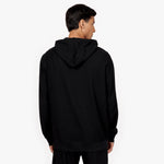 A male model with short, dark hair is shown from the back, standing against a plain white background. He is wearing a black hooded sweatshirt with a relaxed fit. The hood is down, resting flat against his upper back. The back of the hoodie has a clean, minimalistic design with no visible branding or embellishments. He is also wearing matching black sweatpants. His posture is neutral, with his arms resting naturally at his sides, and he is slightly turning his head to the right.