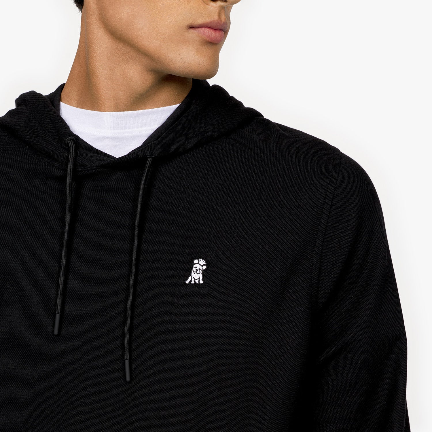 A close-up shot of a male model wearing a black hooded sweatshirt. The fabric has a smooth texture, and the hood features black drawstrings with matching black aglets. A small embroidered logo of a sitting dog in white is visible on the left chest area. The model is also wearing a white crew-neck T-shirt underneath, slightly visible at the neckline. His face is partially shown, revealing his jawline and lips as he looks to the side.