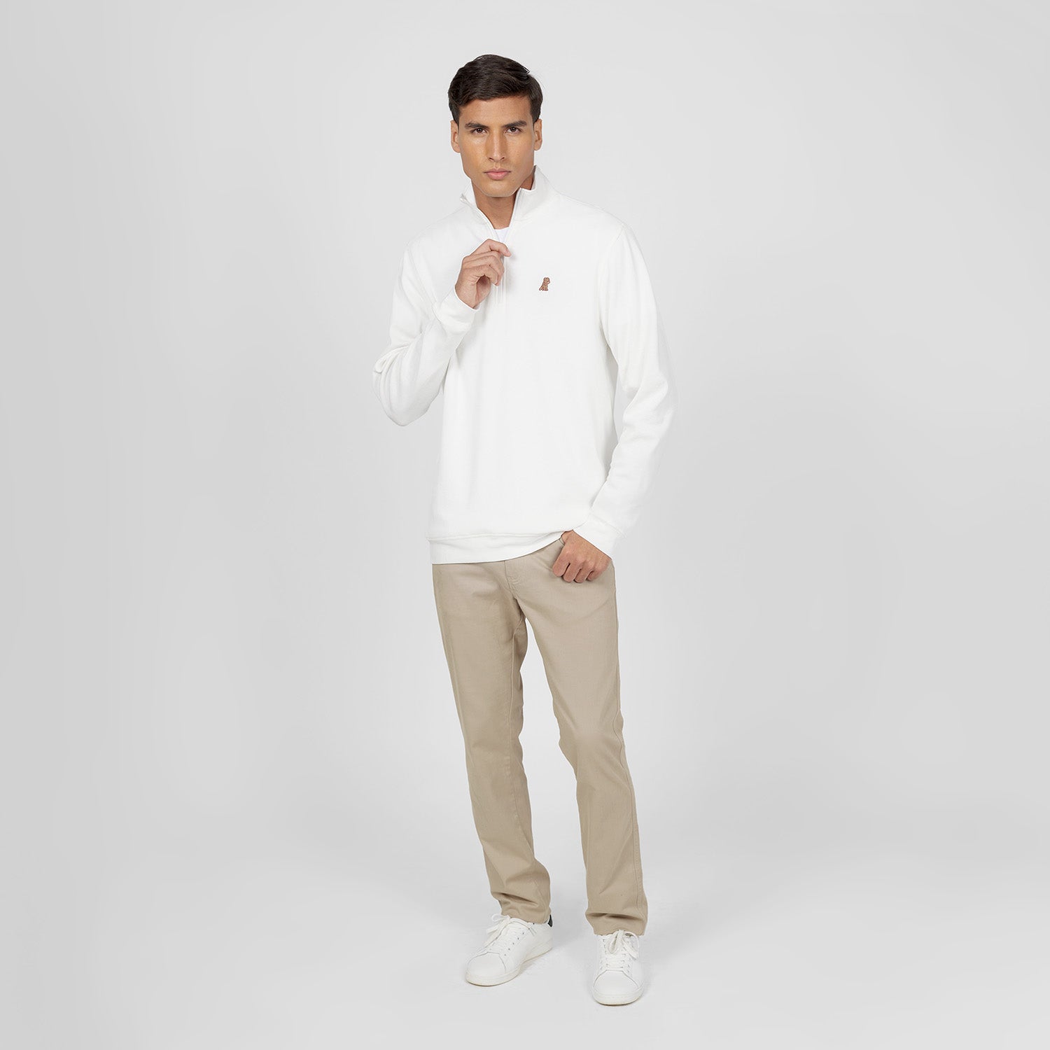 Full-body shot of a man wearing a white half-zip sweatshirt with a small embroidered dog logo on the left chest. He is paired with beige pants and white sneakers. The model is slightly adjusting the zipper with one hand while the other rests in his pocket. The background is plain light gray