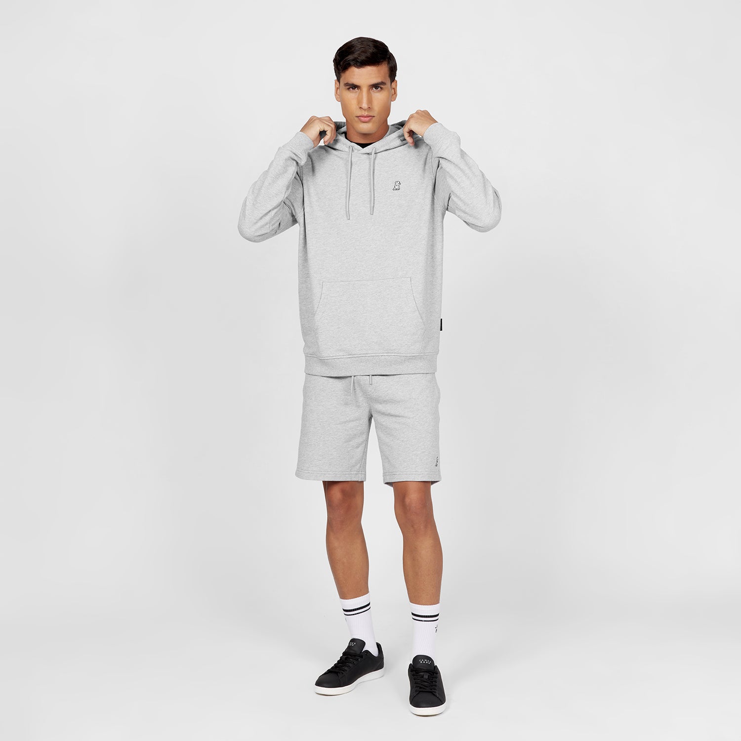 Full-body image of a man wearing a light gray hoodie with a kangaroo pocket, drawstring, and an embroidered dog logo on the chest, paired with matching light gray shorts. He is also wearing white crew socks with black stripes and black sneakers. The man is standing in a casual pose, adjusting the hoodie’s drawstrings, against a plain white background." Let me know if you'd like any changes