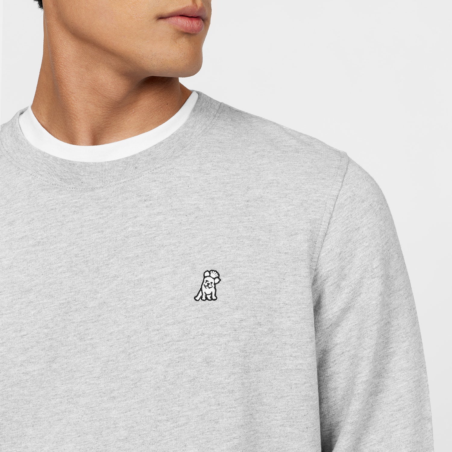 Close-up of a light gray crewneck sweatshirt featuring an embroidered logo of a small, stylized dog on the left chest. The sweatshirt is paired with a white undershirt, highlighting the casual and comfortable design