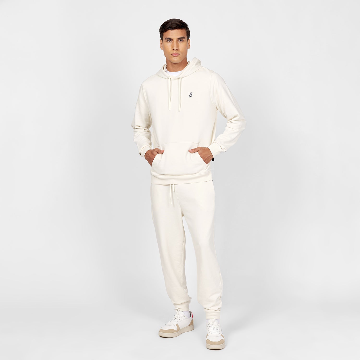 Full-body image of a man wearing an off-white hoodie with a kangaroo pocket, drawstring, and a small embroidered dog logo on the chest. The outfit is paired with matching off-white jogger-style sweatpants and white sneakers with beige soles and pink accents on the heels. The man is standing in a casual pose against a plain white background." Let me know if you'd like to make any changes