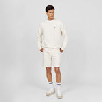 Full-body shot of a man dressed in an off-white crewneck sweatshirt paired with matching off-white shorts. He is wearing white socks with black stripes and white sneakers, styled for a clean and casual look. The embroidered dog logo on the sweatshirt adds a subtle detail to the outfit