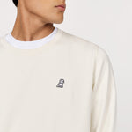 Close-up of a man wearing an off-white crewneck sweatshirt over a white T-shirt. The sweatshirt features a small embroidered logo of a dog on the left chest, adding a subtle yet stylish detail to the minimalist design