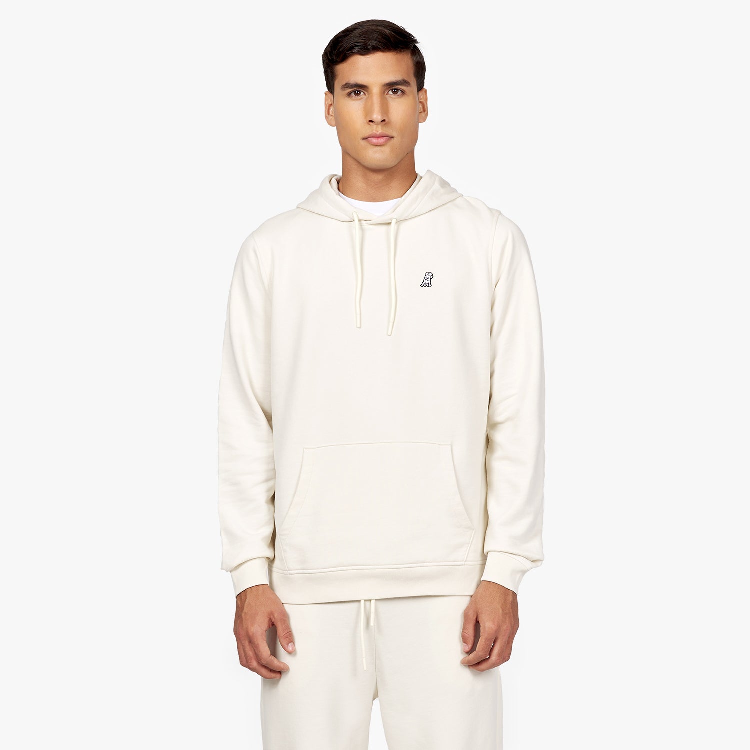 Front view of a man wearing an off-white hoodie with a drawstring and a small embroidered dog logo on the chest. The hoodie features a front kangaroo pocket and is paired with matching off-white sweatpants. The man is standing against a plain white background." Let me know if you'd like any adjustments