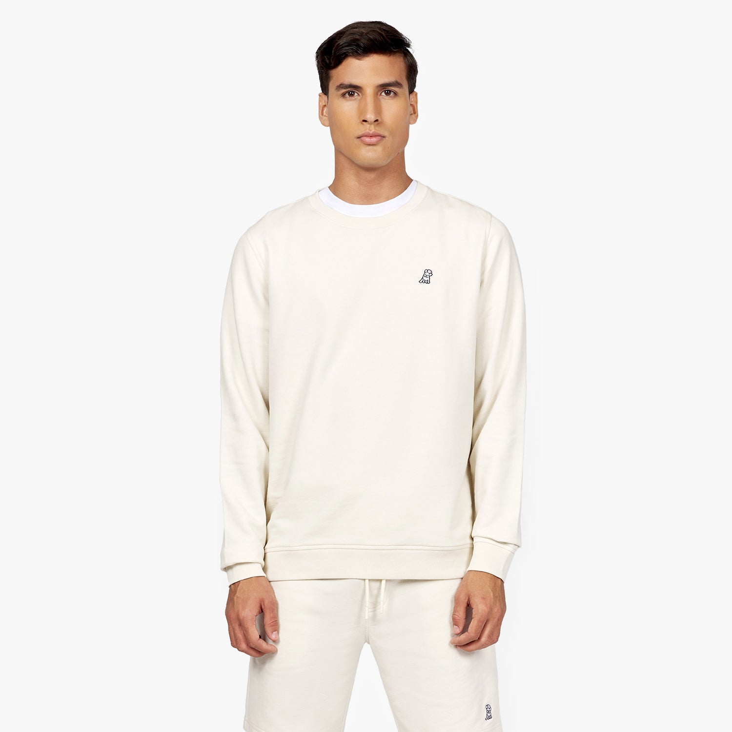 Front-facing view of a man wearing an off-white crewneck sweatshirt with a small embroidered black dog logo on the chest. The sweatshirt is paired with matching off-white sweatpants. The outfit is layered over a white t-shirt for a clean and minimalistic look