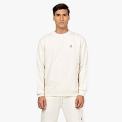 Front-facing view of a man wearing an off-white crewneck sweatshirt with a small embroidered black dog logo on the chest. The sweatshirt is paired with matching off-white sweatpants. The outfit is layered over a white t-shirt for a clean and minimalistic look