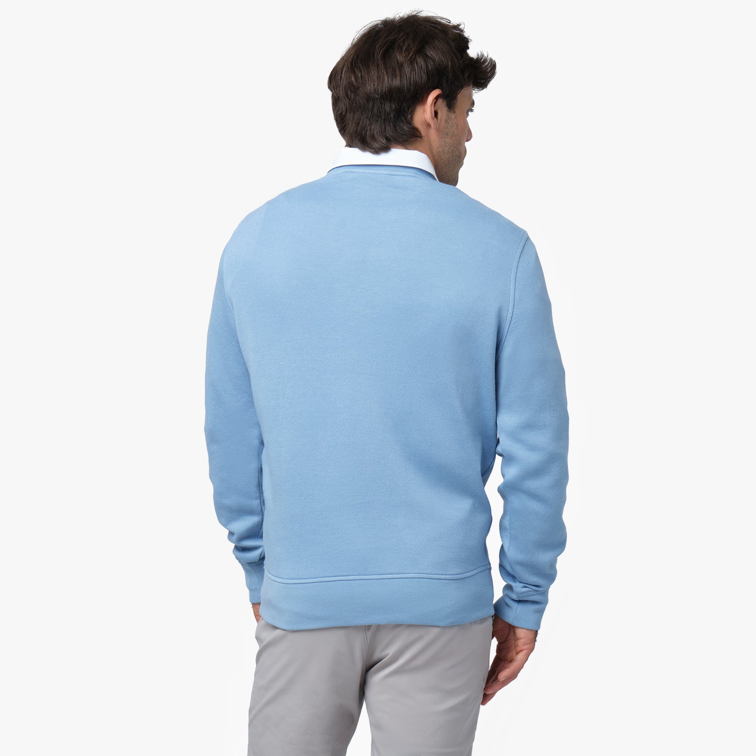 A man with short brown hair and light skin is seen from the back, wearing a light blue crewneck sweatshirt over a white collared shirt. The sweatshirt has a simple design with no visible logos or patterns on the back. He is also wearing light gray pants. He stands against a plain white background, slightly turning his head to the side.