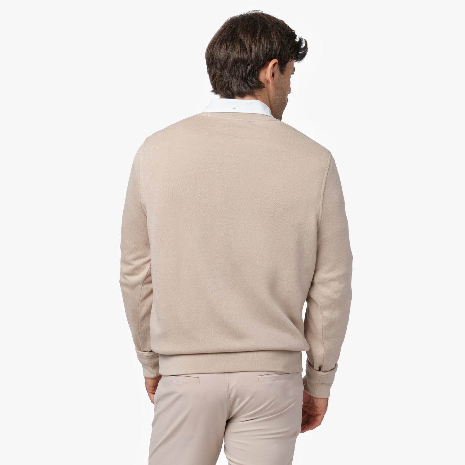 A man with short brown hair and light skin is seen from the back, wearing a beige crewneck sweatshirt over a white collared shirt. The sweatshirt has a simple, clean design with no visible logos or patterns on the back. He is also wearing matching beige pants. He stands against a plain white background, slightly turning his head to the side.