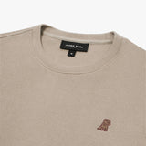 A close-up of a beige crewneck sweatshirt, showing the ribbed collar and finely knit fabric texture. The black neck label displays the brand name "JAMES BARK" with "Est. 2015" beneath it, along with the size "M." A small embroidered logo of a dog in brown thread is positioned on the left chest area, subtly contrasting with the fabric. The material appears soft and high-quality.