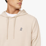 A close-up shot of a male model wearing a beige hooded sweatshirt. The fabric has a smooth texture, and the hood features drawstrings with matching beige aglets. A small embroidered logo of a sitting dog in black is visible on the left chest area. The model is also wearing a white crew-neck T-shirt underneath, slightly visible at the neckline. His face is partially shown, revealing his jawline and lips as he looks to the side.