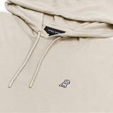 A close-up of a beige hoodie featuring a drawstring hood with matching beige cords and plastic aglets. The fabric has a textured weave, adding a subtle dimension to the design. The chest area displays an embroidered logo of a small white dog wearing a black hat. The brand label, "James Bark," is visible inside the hoodie, stitched onto the neckline.