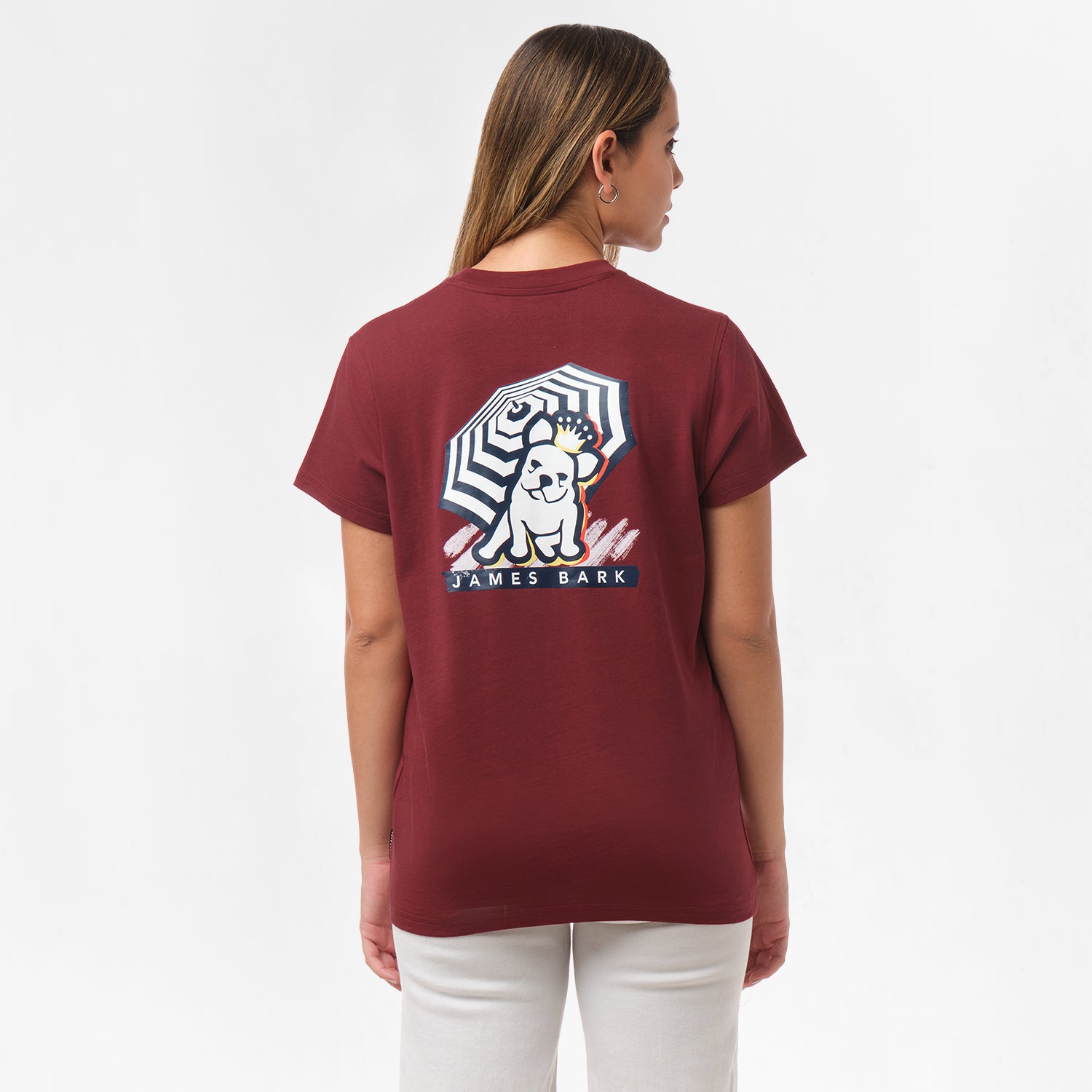 A woman is shown from behind, wearing a maroon T-shirt with a large graphic on the back. The graphic features a dog sitting under a striped umbrella with the text "James Bark" below. She pairs the shirt with white pants.