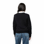 A woman with long, dark hair is shown from the back, wearing a black long-sleeve sweatshirt with ribbed cuffs and hem, layered over a white collared shirt. The sweatshirt has a relaxed fit and a smooth, minimalistic design. She pairs it with classic blue jeans. Her head is slightly turned to the side, showing a partial profile. The background is plain white.