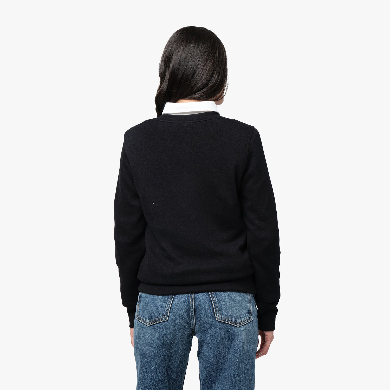 Back view of a woman wearing a black sweater layered over a white collared shirt, paired with blue denim jeans, against a plain white background.