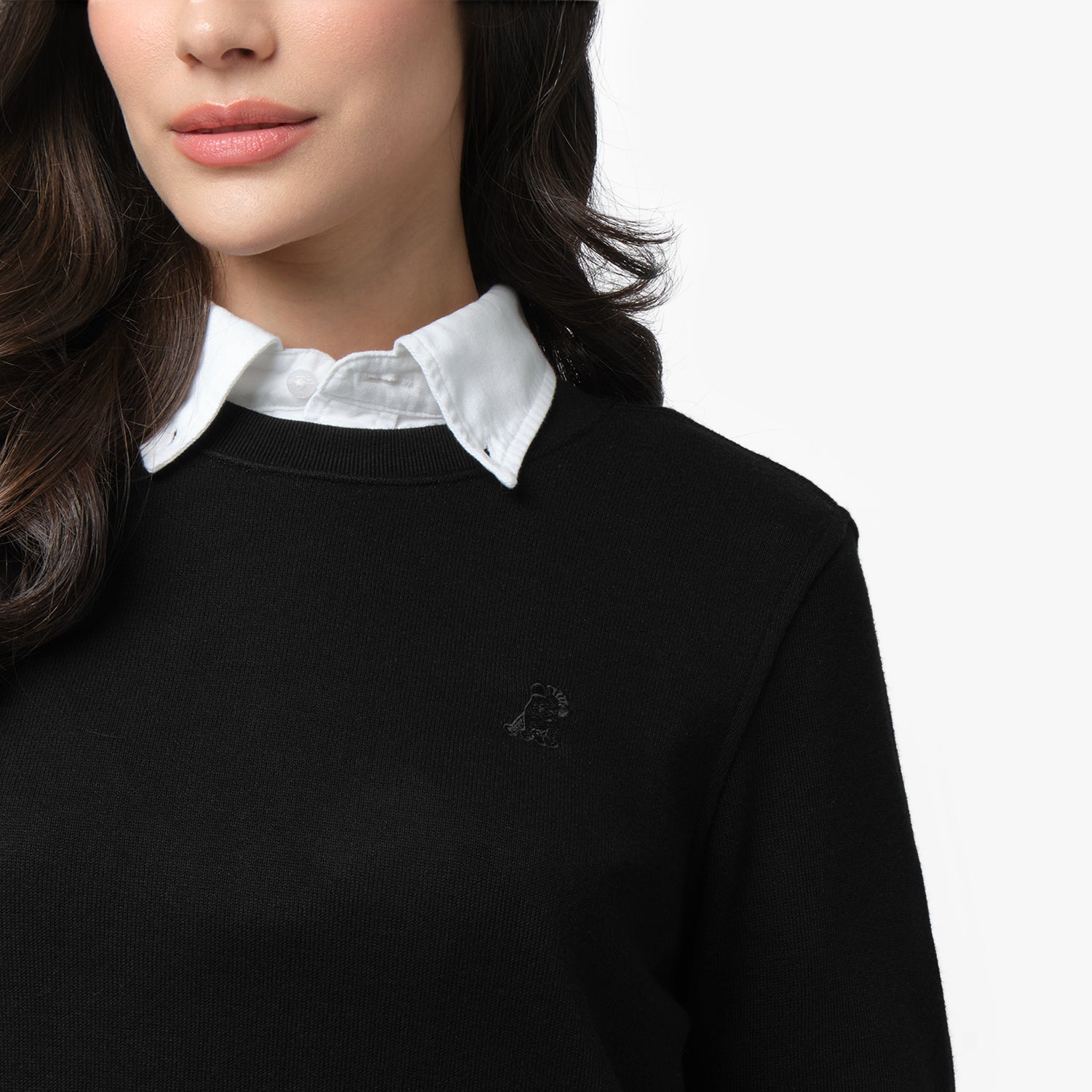 A close-up of a woman wearing a black long-sleeve sweatshirt layered over a crisp white collared shirt. The sweatshirt features a subtle black-on-black embroidered dog logo on the left chest. The ribbed crew neckline sits neatly over the shirt collar. Part of her long, wavy dark hair is visible as she looks slightly to the side. The background is plain white.