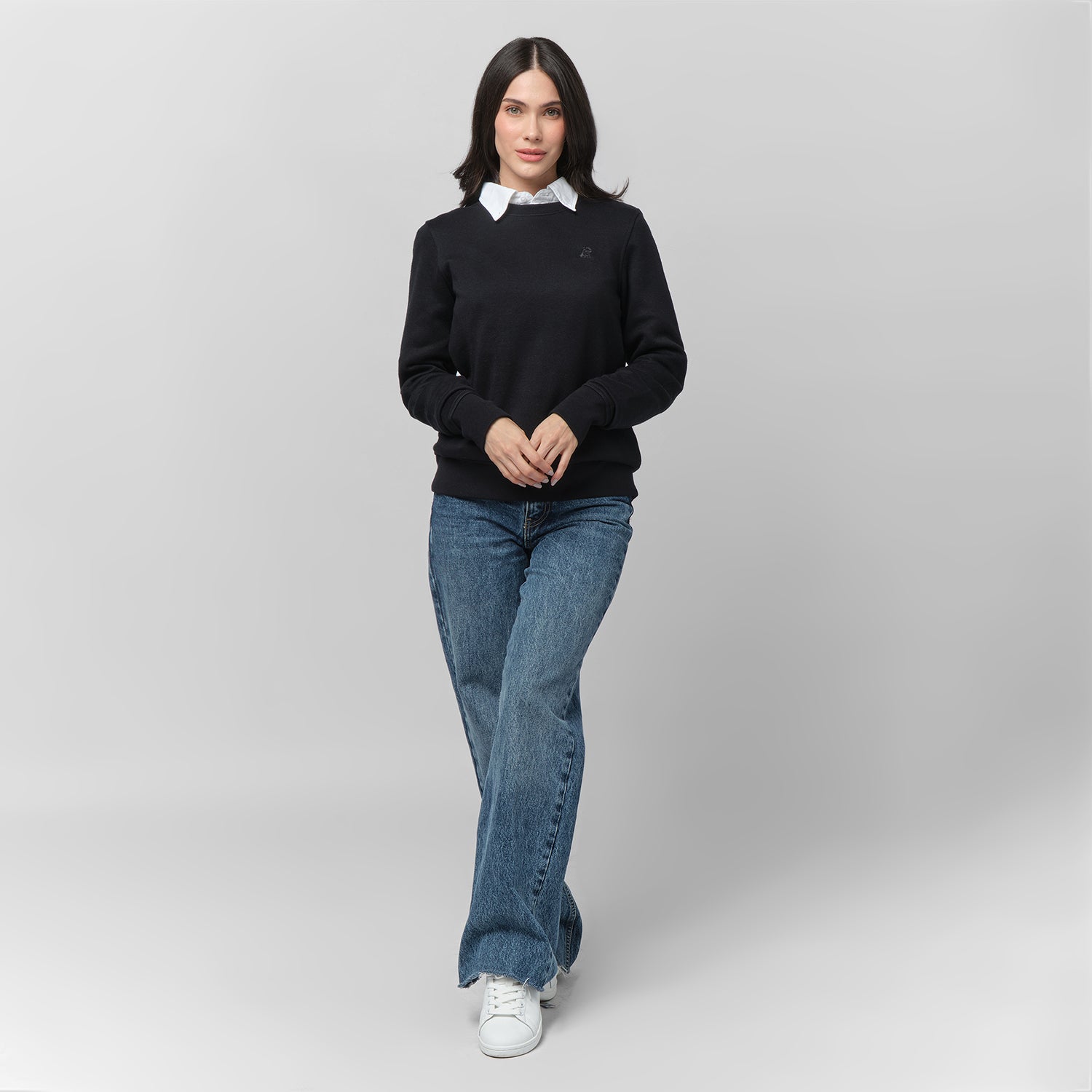 A woman with shoulder-length dark hair walks forward with a composed expression, wearing a black long-sleeve sweatshirt layered over a white collared shirt. The sweatshirt has ribbed cuffs and hem, along with a subtle black-on-black embroidered dog logo on the left chest. She pairs it with classic blue jeans and white sneakers. The background is light gray.