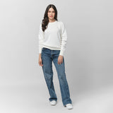 A woman with long, wavy dark hair stands with a relaxed posture, wearing a white long-sleeve sweatshirt with ribbed cuffs and hem. The sweatshirt has a small, subtle embroidered logo on the left chest. She pairs it with high-waisted, slightly flared blue jeans and white sneakers. She looks directly at the camera with a gentle expression. The background is light gray.