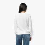 A woman with long, dark hair is shown from the back, wearing a white long-sleeve sweatshirt with a ribbed hem and cuffs. The sweatshirt has a clean, minimalistic design with a relaxed fit. She pairs it with classic blue jeans. Her head is slightly turned to the side, showing a partial profile. The background is plain white.