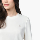 A close-up of a woman wearing a white long-sleeve sweatshirt with a ribbed crew neckline that has a subtle beige trim. The sweatshirt features a small embroidered dog logo on the left chest in a matching beige tone. Part of her long, wavy dark hair is visible as she looks slightly to the side. The background is plain white.