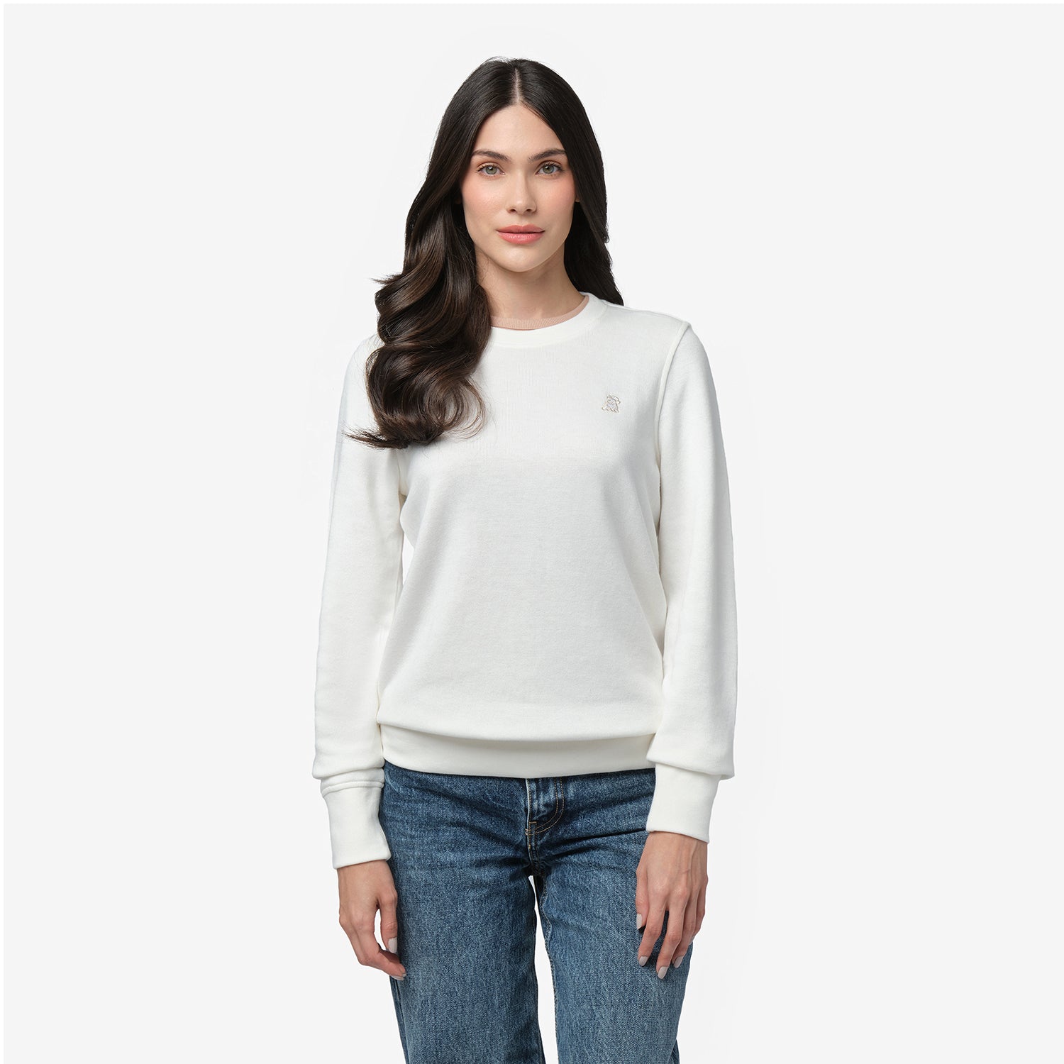 A woman with long, wavy dark hair and a gentle smile wears a white long-sleeve sweatshirt with a ribbed crew neckline and cuffs. The sweatshirt features a small, subtle embroidered logo on the left chest. She pairs it with classic blue jeans. She stands against a plain white background, looking directly at the camera with a relaxed posture.