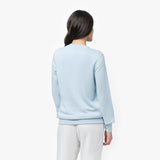  A woman with long, dark hair is shown from the back, wearing a light blue long-sleeve sweatshirt with ribbed cuffs and hem. The sweatshirt has a relaxed fit and a smooth, minimalistic design. She pairs it with white wide-leg pants. Her head is slightly turned to the side, showing a partial profile. The background is plain white