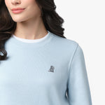 A close-up of a woman wearing a light blue long-sleeve sweatshirt with a ribbed crew neckline layered over a white undershirt. The sweatshirt features a small embroidered dog logo on the left chest in a darker blue tone. Part of her long, wavy dark hair is visible as she looks slightly to the side. The background is plain white.