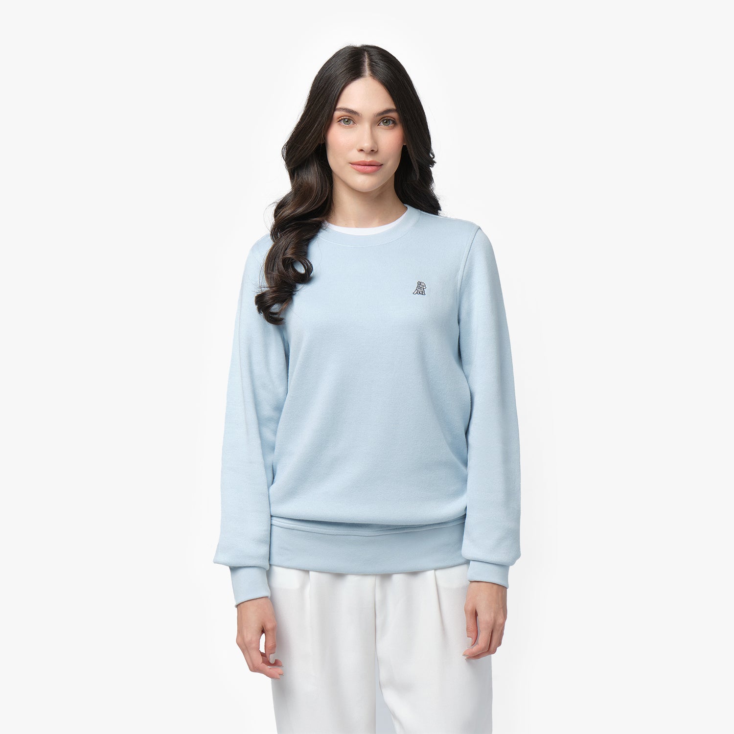 A woman with long, wavy dark hair and a calm expression wears a light blue long-sleeve sweatshirt with a ribbed crew neckline, cuffs, and hem. The sweatshirt features a small embroidered dog logo on the left chest. She pairs it with white wide-leg pants. She stands against a plain white background, looking directly at the camera with a relaxed posture.