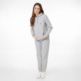 Full-body image of a woman wearing a light gray hoodie with a small embroidered dog logo, matching sweatpants, and white sneakers. She has long brown hair, gold hoop earrings, and is standing in a relaxed pose with hands in the hoodie pocket, against a plain white background." Let me know if you'd like modifications