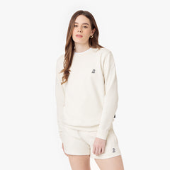 Woman wearing an off-white crewneck sweatshirt and matching shorts, featuring a subtle embroidered logo on the chest and shorts. Styled with gold hoop earrings and a relaxed, casual look.