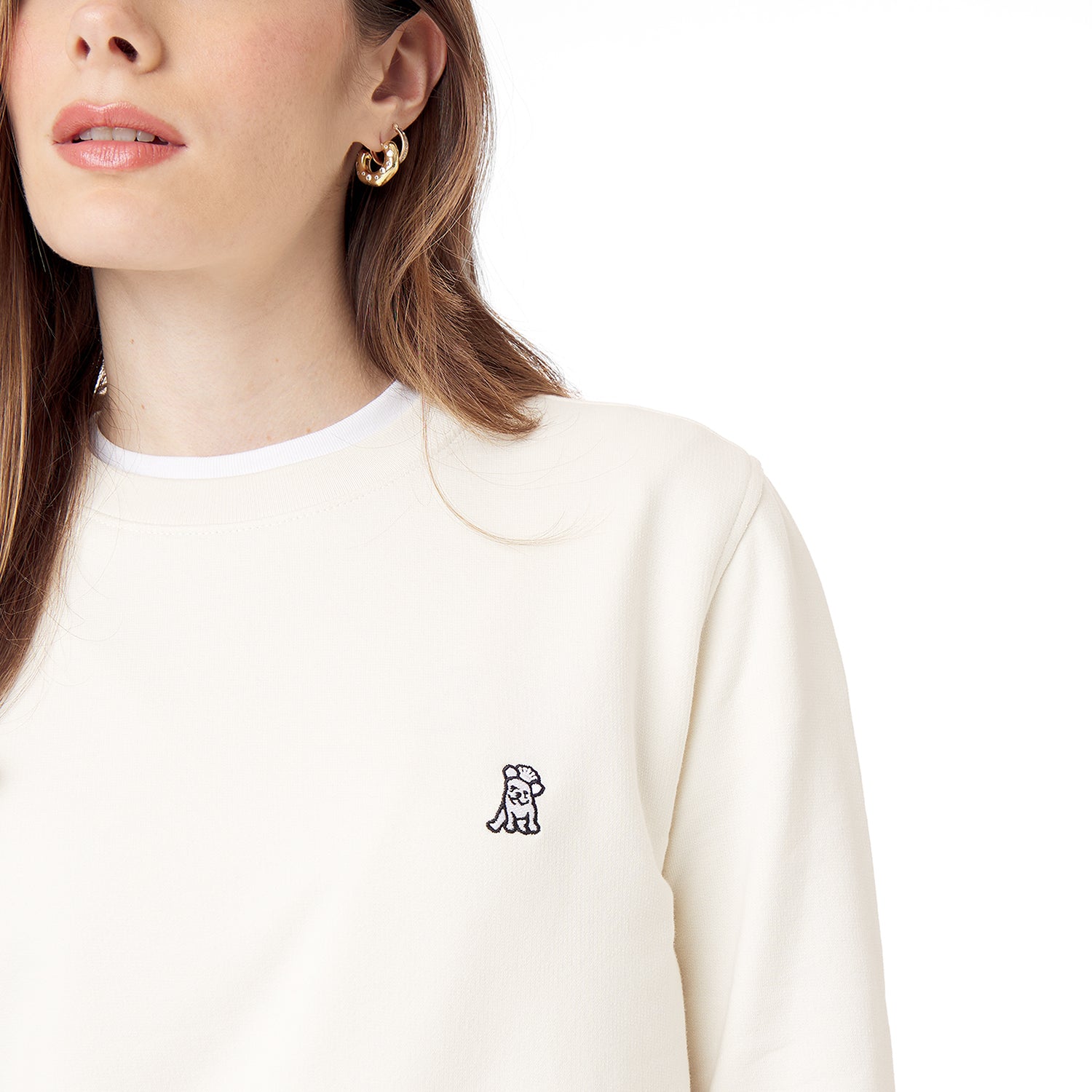 Close-up of a woman wearing an off-white crewneck sweatshirt, highlighting the embroidered French Bulldog logo on the chest. The minimalist design complements her gold hoop earrings.