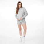 Woman wearing a light gray sweatshirt and matching shorts, styled with white sneakers and striped socks. She has long wavy hair and is standing in a casual pose against a white background.