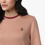 A close-up of a woman wearing a dusty pink long-sleeve sweatshirt with a ribbed crew neckline featuring a contrasting burgundy trim. The sweatshirt has a small embroidered dog logo on the left chest in a matching brown tone. Part of her long, wavy dark hair is visible as she looks slightly to the side. The background is plain white