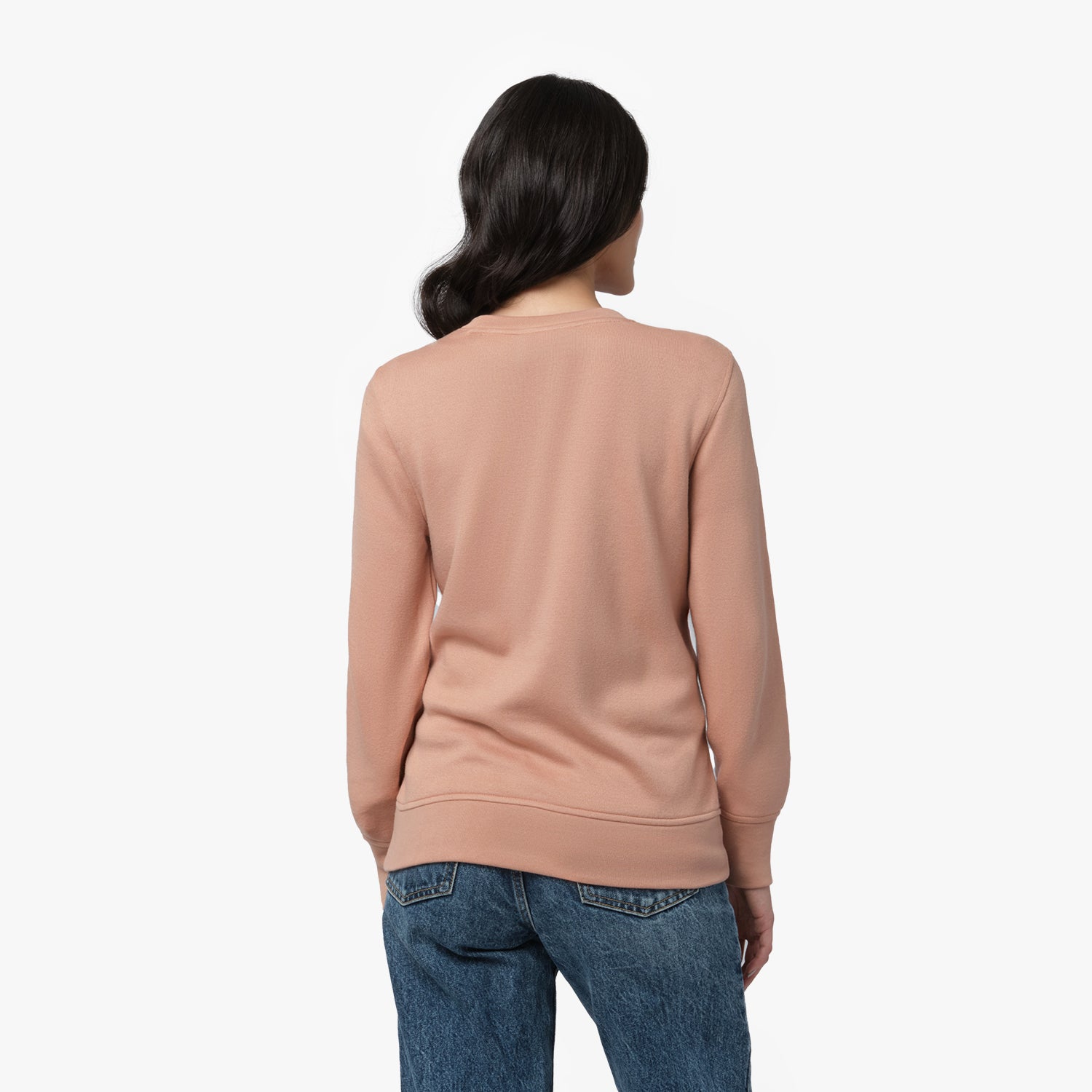 A woman with long, dark hair is shown from the back, wearing a dusty pink long-sleeve sweatshirt with ribbed cuffs and hem. The sweatshirt has a relaxed fit and a smooth, minimalistic design. She pairs it with classic blue jeans. Her head is slightly turned to the side, showing a partial profile. The background is plain white.