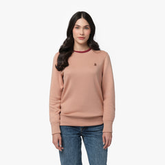 A woman with long, wavy dark hair and a calm expression wears a dusty pink long-sleeve sweatshirt with ribbed cuffs and hem. The sweatshirt features a small embroidered dog logo on the left chest and has a contrasting burgundy trim around the crew neckline. She pairs it with classic blue jeans. She stands against a plain white background, looking directly at the camera with a relaxed posture.