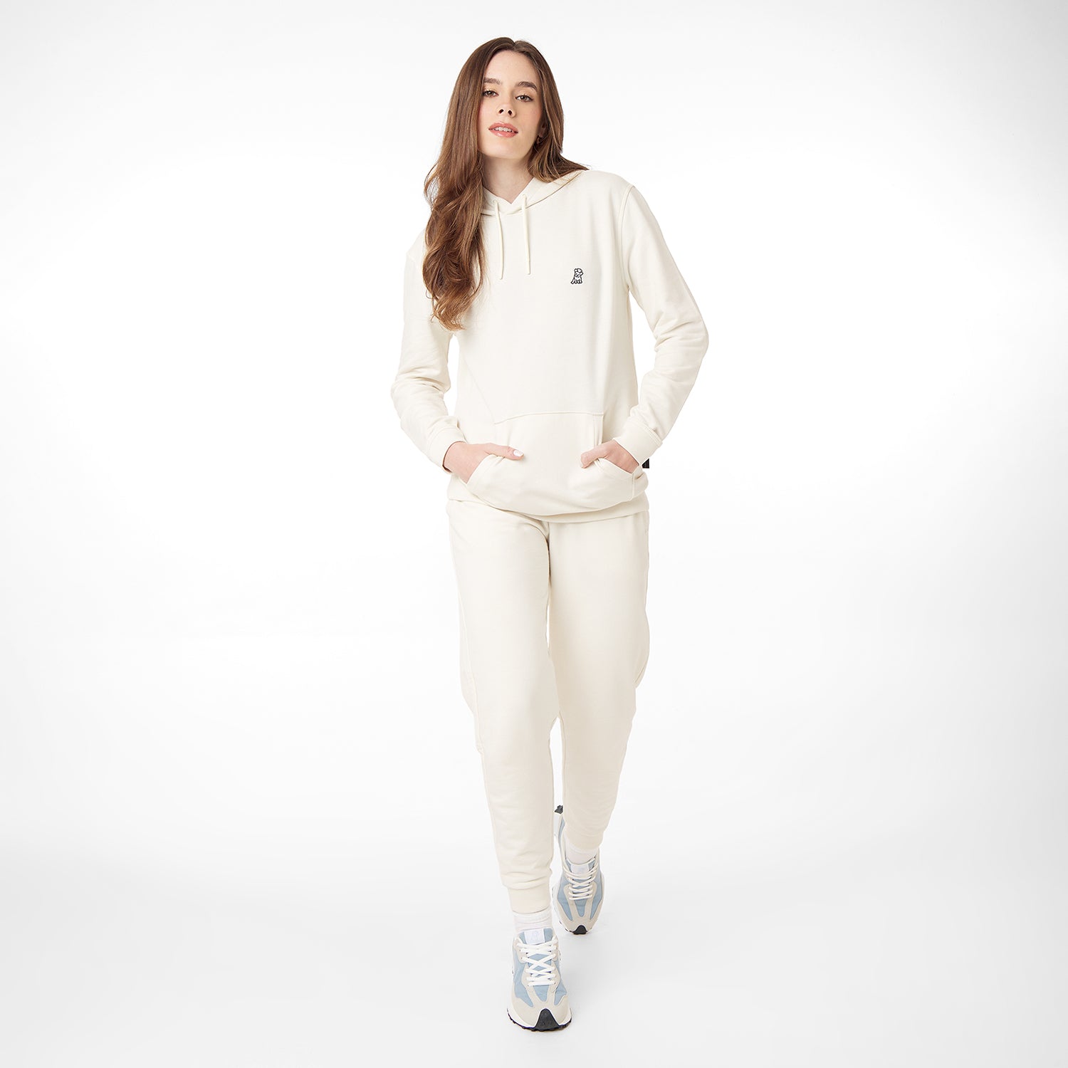 A woman wearing a cream-colored hoodie with a subtle embroidered Frenchie design, paired with matching joggers and blue-and-white sneakers, posing in a walking stance against a white background.