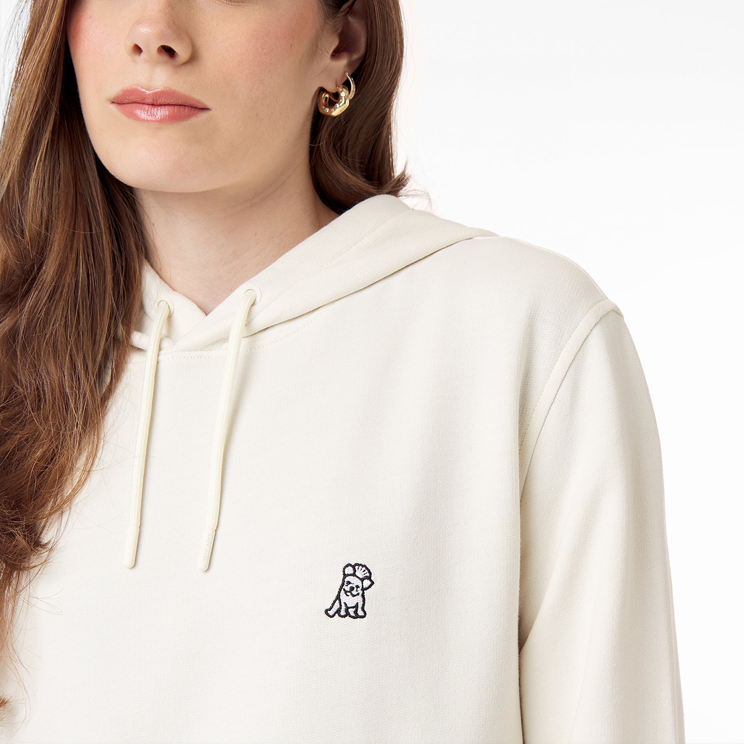 Close-up of a woman wearing a cream-colored hoodie featuring a subtle embroidered Frenchie design on the chest, paired with gold hoop earrings.