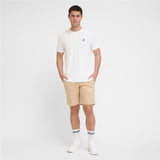 A man modeling a white T-shirt with a small black logo on the left chest, paired with beige shorts, white socks, and white sneakers