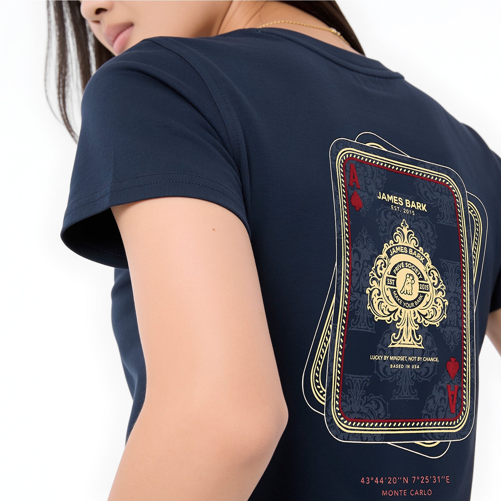 A close-up of a model wearing a navy blue T-shirt, showing the back graphic. The graphic is styled like a playing card deck with gold and red details, featuring the text 'JAMES BARK EST'.