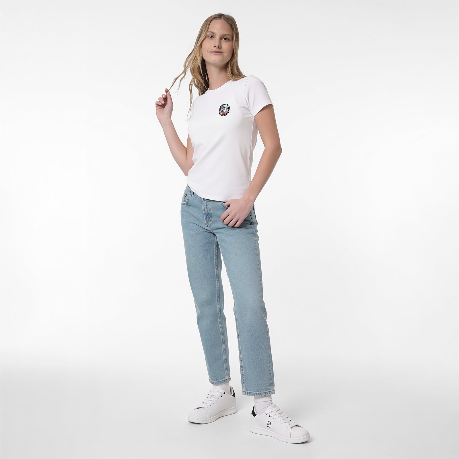 A woman modeling a white T-shirt with a small circular graphic on the left chest, paired with light-wash blue jeans