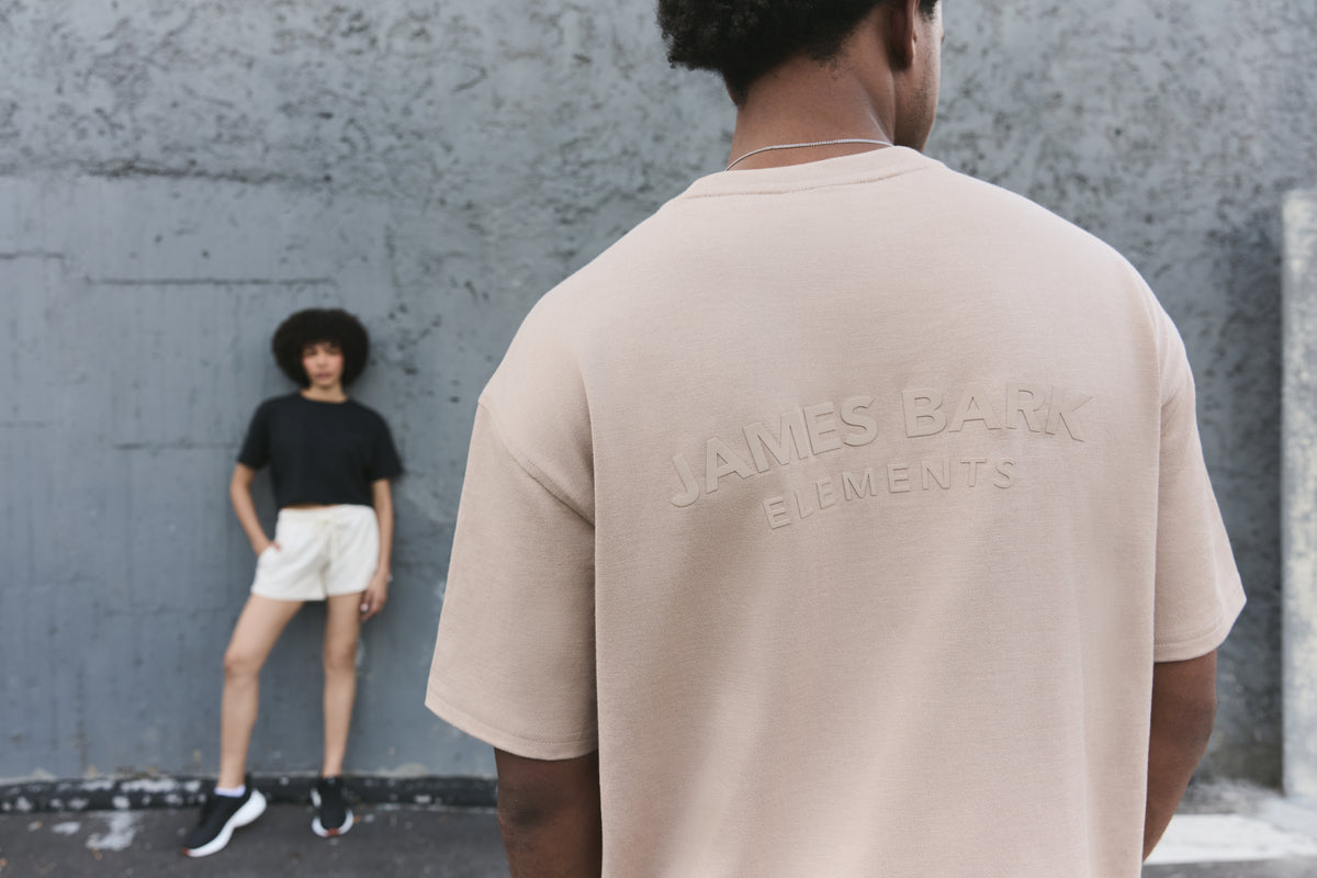 A person shown from behind wearing an oversized light beige t-shirt, with a model in a black t-shirt and white shorts standing in the background