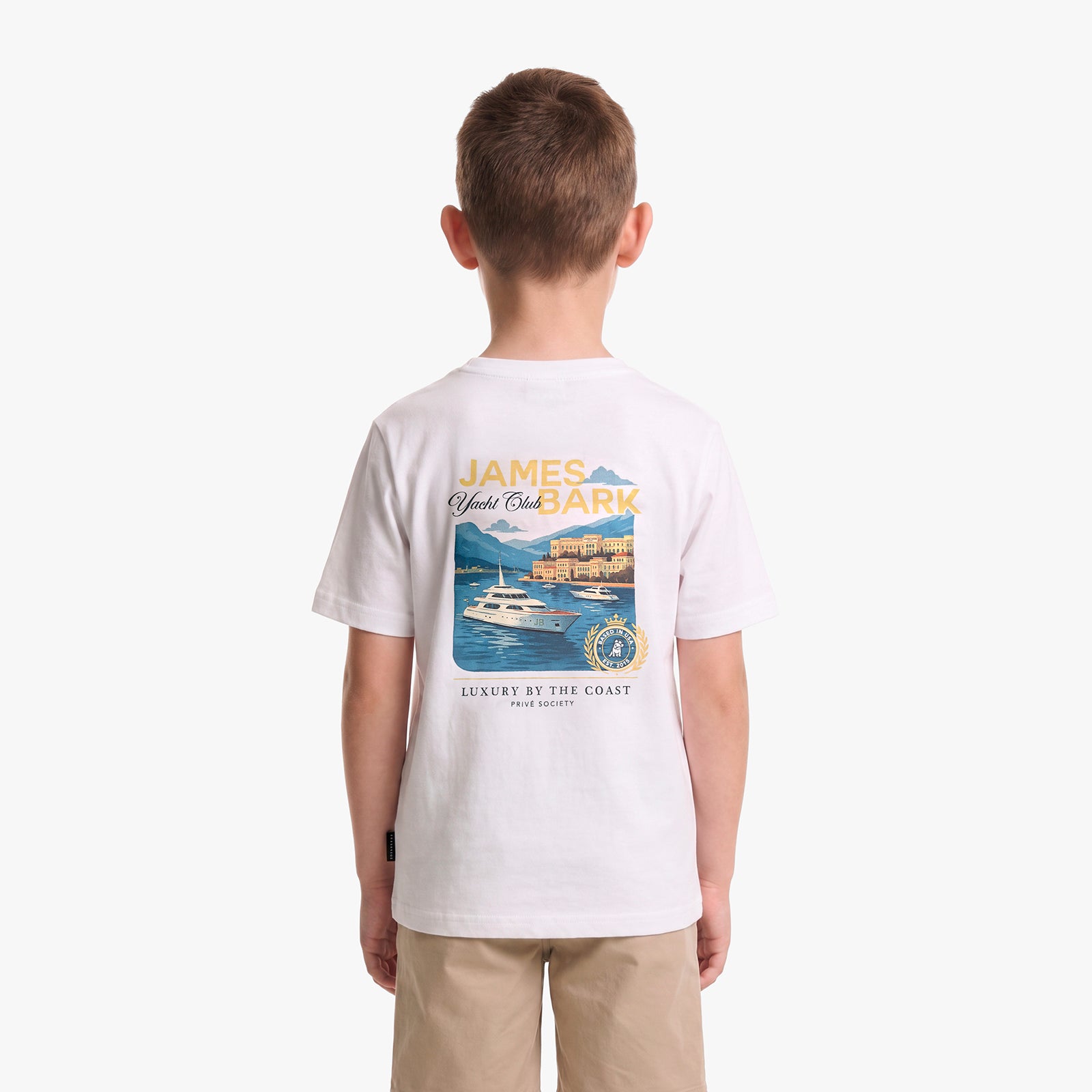 A child viewed from behind, wearing a white T-shirt with a large multicolor graphic print on the back featuring a yacht and coastal scene
