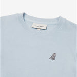 A close-up of a light pastel blue crewneck sweatshirt, showing the ribbed collar and finely knit fabric texture. The white neck label displays the brand name "JAMES BARK" with "Est. 2015" beneath it. A small embroidered logo of a dog in dark thread is positioned on the left chest area, standing out against the light fabric. The material appears soft and high-qua